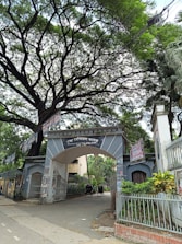An entrance archway to Dhaka Medical College is surrounded by lush greenery and covered with several posters. There are trees overhead with sprawling branches, creating a canopy. Two rickshaws are parked inside the gate, indicative of a busy, urban setting.