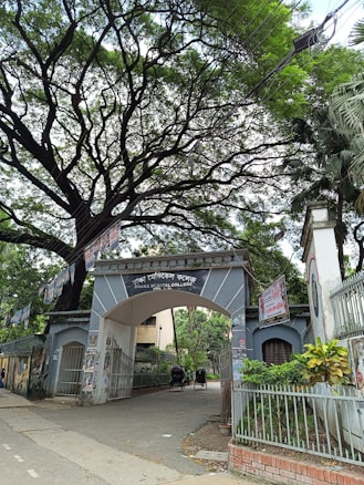 An entrance archway to Dhaka Medical College is surrounded by lush greenery and covered with several posters. There are trees overhead with sprawling branches, creating a canopy. Two rickshaws are parked inside the gate, indicative of a busy, urban setting.