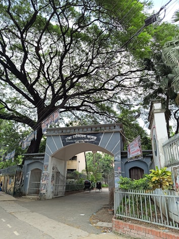 An entrance archway to Dhaka Medical College is surrounded by lush greenery and covered with several posters. There are trees overhead with sprawling branches, creating a canopy. Two rickshaws are parked inside the gate, indicative of a busy, urban setting.