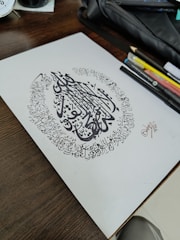 A piece of paper with intricate Arabic calligraphy surrounded by artistic writing in a circular pattern. The paper rests on a wooden table alongside various pens, pencils, and a black pouch.