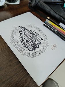 A piece of paper with intricate Arabic calligraphy surrounded by artistic writing in a circular pattern. The paper rests on a wooden table alongside various pens, pencils, and a black pouch.