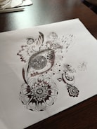 Detailed pen art featuring intricate patterns and designs on a sheet of paper, with the drawing emphasizing circular and swirl motifs. The artwork rests on a dark wooden table surface with some stationary items visible in the background.