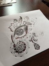 Detailed pen art featuring intricate patterns and designs on a sheet of paper, with the drawing emphasizing circular and swirl motifs. The artwork rests on a dark wooden table surface with some stationary items visible in the background.
