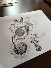 Detailed pen art featuring intricate patterns and designs on a sheet of paper, with the drawing emphasizing circular and swirl motifs. The artwork rests on a dark wooden table surface with some stationary items visible in the background.