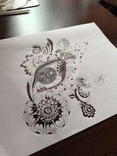 Detailed pen art featuring intricate patterns and designs on a sheet of paper, with the drawing emphasizing circular and swirl motifs. The artwork rests on a dark wooden table surface with some stationary items visible in the background.
