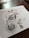 Detailed pen art featuring intricate patterns and designs on a sheet of paper, with the drawing emphasizing circular and swirl motifs. The artwork rests on a dark wooden table surface with some stationary items visible in the background.