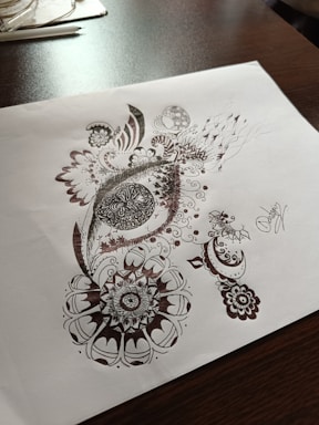Detailed pen art featuring intricate patterns and designs on a sheet of paper, with the drawing emphasizing circular and swirl motifs. The artwork rests on a dark wooden table surface with some stationary items visible in the background.