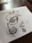 Detailed pen art featuring intricate patterns and designs on a sheet of paper, with the drawing emphasizing circular and swirl motifs. The artwork rests on a dark wooden table surface with some stationary items visible in the background.