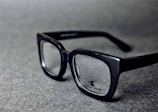 A pair of black rectangular eyeglasses with clear lenses sits on a grey textured surface. The glasses have a glossy finish and the brand logo is visible on one of the lenses.