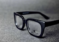 A pair of black rectangular eyeglasses with clear lenses sits on a grey textured surface. The glasses have a glossy finish and the brand logo is visible on one of the lenses.