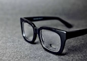 A pair of black rectangular eyeglasses with clear lenses sits on a grey textured surface. The glasses have a glossy finish and the brand logo is visible on one of the lenses.