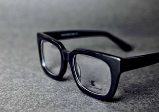 A pair of black rectangular eyeglasses with clear lenses sits on a grey textured surface. The glasses have a glossy finish and the brand logo is visible on one of the lenses.