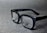 A pair of black rectangular eyeglasses with clear lenses sits on a grey textured surface. The glasses have a glossy finish and the brand logo is visible on one of the lenses.