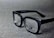 A pair of black rectangular eyeglasses with clear lenses sits on a grey textured surface. The glasses have a glossy finish and the brand logo is visible on one of the lenses.