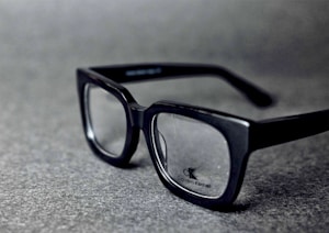 A pair of black rectangular eyeglasses with clear lenses sits on a grey textured surface. The glasses have a glossy finish and the brand logo is visible on one of the lenses.