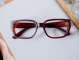 A pair of rectangular, brown eyeglasses rests on a white surface. The frame is thick with a gradient that fades into a lighter brown. Part of a wooden picture frame is visible at the corner, hinting at a desk setting.