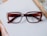 A pair of rectangular, brown eyeglasses rests on a white surface. The frame is thick with a gradient that fades into a lighter brown. Part of a wooden picture frame is visible at the corner, hinting at a desk setting.