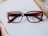 A pair of rectangular, brown eyeglasses rests on a white surface. The frame is thick with a gradient that fades into a lighter brown. Part of a wooden picture frame is visible at the corner, hinting at a desk setting.
