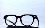 Black-framed eyeglasses placed on a dark surface with a light, minimalistic background. The glasses have a classic design with clear lenses.