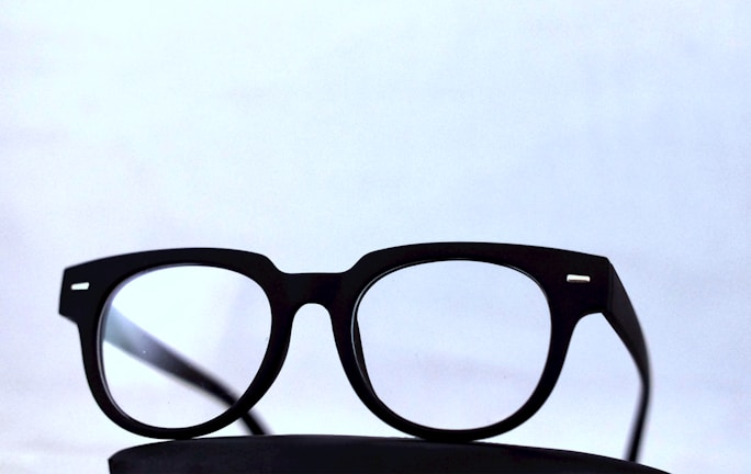 Black-framed eyeglasses placed on a dark surface with a light, minimalistic background. The glasses have a classic design with clear lenses.