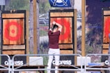 A person is standing in front of a wooden target, preparing to throw an axe. The target has a red bullseye painted on it. The scene is part of an outdoor axe-throwing area, enclosed by netting with multiple targets visible.