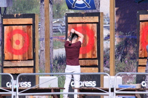 A person is standing in front of a wooden target, preparing to throw an axe. The target has a red bullseye painted on it. The scene is part of an outdoor axe-throwing area, enclosed by netting with multiple targets visible.