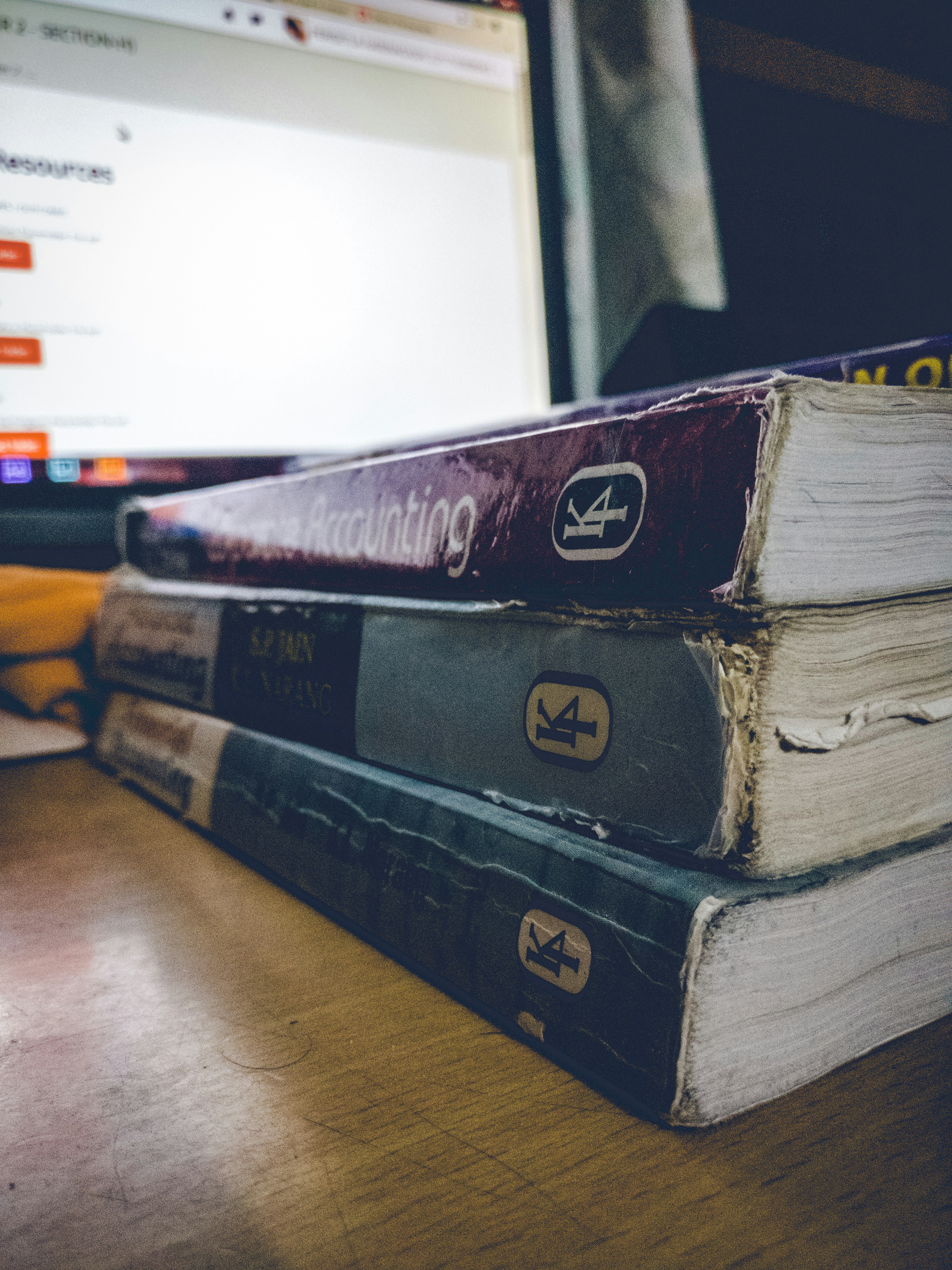 A stack of three worn textbooks with visible spines, labeled with different subjects, sits on a wooden table beside a glowing laptop screen displaying a website. The focus is on the books, with their aged and rough edges noticeable.