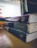 A stack of three worn textbooks with visible spines, labeled with different subjects, sits on a wooden table beside a glowing laptop screen displaying a website. The focus is on the books, with their aged and rough edges noticeable.