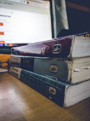 A stack of three worn textbooks with visible spines, labeled with different subjects, sits on a wooden table beside a glowing laptop screen displaying a website. The focus is on the books, with their aged and rough edges noticeable.