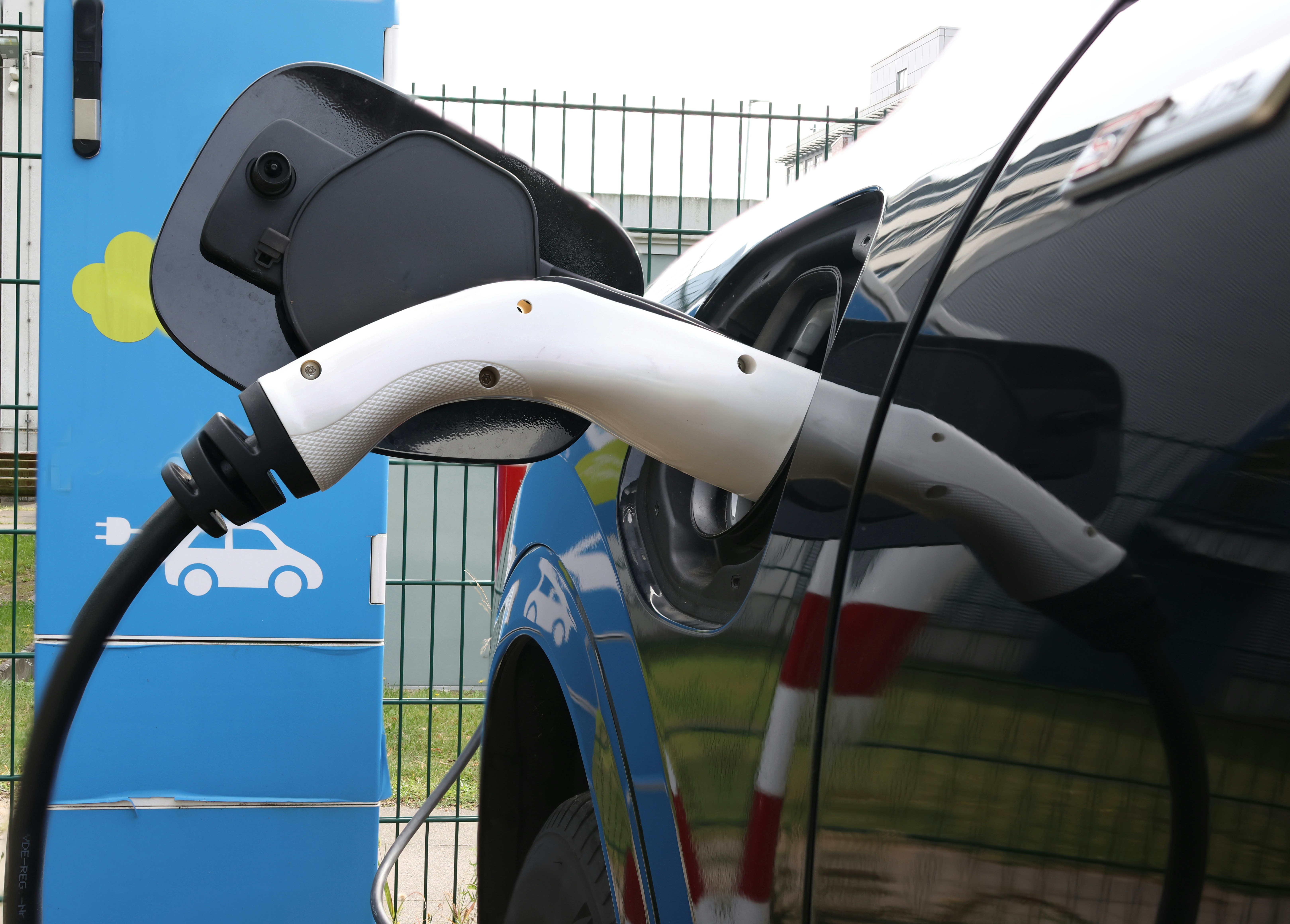An electric vehicle is connected to a charging station. The charging cable is plugged into the car's charging port. The station has a blue and green color scheme with icons indicating its purpose. The scene is set outdoors with a metal fence in the background.