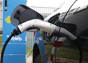An electric vehicle is connected to a charging station. The charging cable is plugged into the car's charging port. The station has a blue and green color scheme with icons indicating its purpose. The scene is set outdoors with a metal fence in the background.