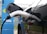 An electric vehicle is connected to a charging station. The charging cable is plugged into the car's charging port. The station has a blue and green color scheme with icons indicating its purpose. The scene is set outdoors with a metal fence in the background.