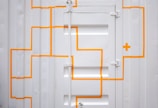 A white corrugated metal door with bright orange, geometric lines and a cross pattern. The door has visible hinges and panels, creating a textured and industrial appearance.