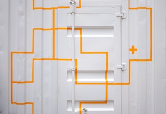 A white corrugated metal door with bright orange, geometric lines and a cross pattern. The door has visible hinges and panels, creating a textured and industrial appearance.