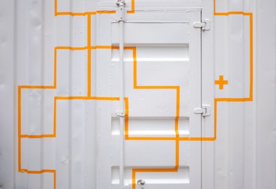 A white corrugated metal door with bright orange, geometric lines and a cross pattern. The door has visible hinges and panels, creating a textured and industrial appearance.