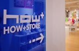 A blue glass panel with the words 'HOW+ STORE' and an arrow pointing to the right. The background contains a bright, well-lit interior of a store with various colorful items displayed.