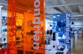 A contemporary retail space with transparent colored panels featuring the names of modern artists and designers. The orange and blue panels provide a vibrant contrast against the showroom, which is well-lit and filled with various design objects and furniture pieces.