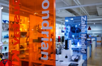 A contemporary retail space with transparent colored panels featuring the names of modern artists and designers. The orange and blue panels provide a vibrant contrast against the showroom, which is well-lit and filled with various design objects and furniture pieces.