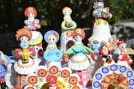 A collection of colorful ceramic figurines representing traditional Eastern European folk characters. The figures are adorned in vibrant costumes featuring floral patterns, and some are holding small animals. They are displayed outdoors on a table with a natural background.