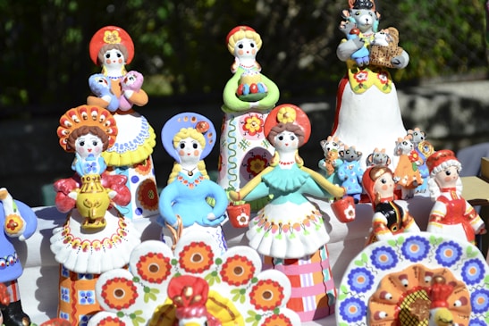A collection of colorful ceramic figurines representing traditional Eastern European folk characters. The figures are adorned in vibrant costumes featuring floral patterns, and some are holding small animals. They are displayed outdoors on a table with a natural background.