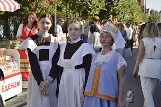 Three women are in traditional clothing, standing outdoors. Two young women wear black dresses with white aprons, resembling maid uniforms, and the older woman wears a blue dress with ornate designs and a decorative headdress. They are part of a larger outdoor gathering or festival, with other people and display stands visible in the background.