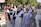 Three women are in traditional clothing, standing outdoors. Two young women wear black dresses with white aprons, resembling maid uniforms, and the older woman wears a blue dress with ornate designs and a decorative headdress. They are part of a larger outdoor gathering or festival, with other people and display stands visible in the background.