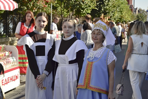 Three women are in traditional clothing, standing outdoors. Two young women wear black dresses with white aprons, resembling maid uniforms, and the older woman wears a blue dress with ornate designs and a decorative headdress. They are part of a larger outdoor gathering or festival, with other people and display stands visible in the background.