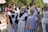 Three women are in traditional clothing, standing outdoors. Two young women wear black dresses with white aprons, resembling maid uniforms, and the older woman wears a blue dress with ornate designs and a decorative headdress. They are part of a larger outdoor gathering or festival, with other people and display stands visible in the background.