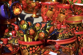 A collection of traditional Russian wooden kitchenware painted with intricate floral designs in bright colors. The items include ladles and pots decorated in red, gold, and black hues, featuring motifs of fruit and leaves.