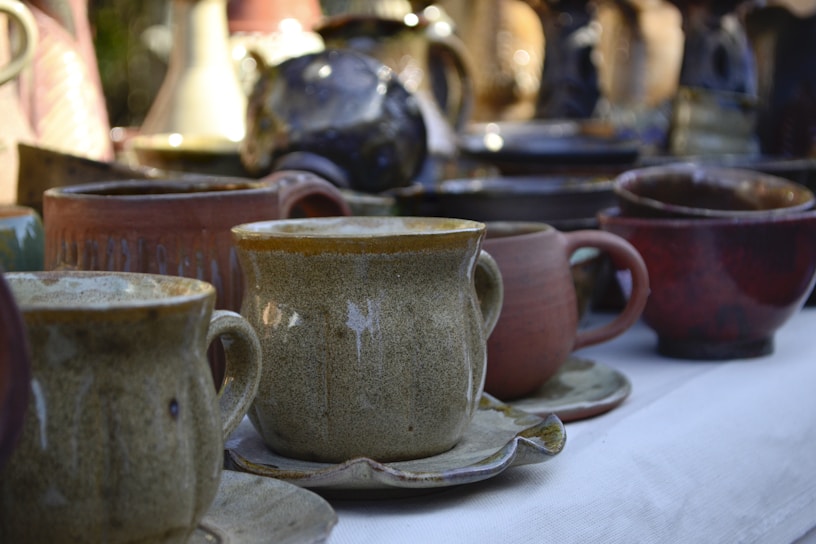 A collection of ceramic mugs and cups of various colors and sizes placed on a fabric surface. The focus is on a central beige mug with a saucer, surrounded by other pottery in shades of brown, red, and blue. The background features a blurred assortment of pottery items.