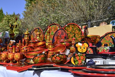 A collection of traditional, hand-painted Russian wooden items featuring ornate floral and fruit designs in vibrant red, black, yellow, and green colors. The items include bowls, plates, and containers, all intricately decorated with folk art patterns. They are displayed outdoors on a white cloth under bright sunlight.