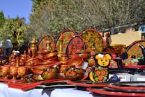 A collection of traditional, hand-painted Russian wooden items featuring ornate floral and fruit designs in vibrant red, black, yellow, and green colors. The items include bowls, plates, and containers, all intricately decorated with folk art patterns. They are displayed outdoors on a white cloth under bright sunlight.