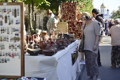 A bustling outdoor market scene with people browsing various stalls displaying crafts and pottery. A woman wearing a sun hat examines items closely, surrounded by an array of earthenware and jewelry. The atmosphere is lively and vibrant, with a mix of shoppers and onlookers. Trees line the street, providing shade on a sunny day, and a church with golden domes is visible in the background.