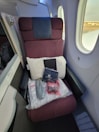 A first-class airplane seat with a maroon fabric upholstery is adjacent to two airplane windows. On the seat, a white pillow, a blanket in clear packaging, a pair of slippers, an amenity kit labeled with 'Japan Airlines', and a small red bag are neatly arranged.